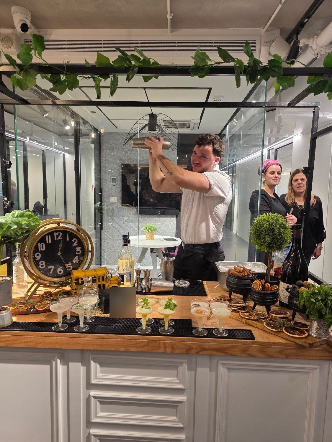 ברמן מנער שייקר מול אורחות, ועל הבר כוסות קוקטייל מוכנות ושעון זהב